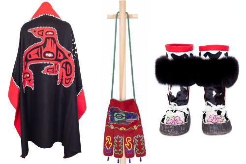 Three objects side by side on white background: a cloak-like blanket with red edges and central design on black. A red bag with long blue strap and animal design in blue, hanging from a wooden rack. A pair of booths with black furry fringe, pink blossom e