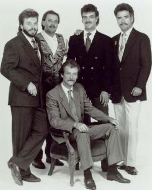 Four men in suits pose behind a fifth man sitting down in a promotional band photo. Black-and-white photo.
