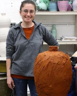 A person poses next to the reddish-brown clay jug, which is about the size of their torso and matches the color of their T-shirt.