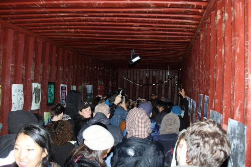A crowd of people cram inside a sea container, with red walls and ceiling, and art prints affixed to the walls.