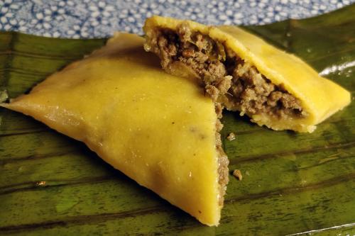 Close-up of meat-filled hand pie with yellow crust, cut in half on the diagonal, resting on top of a dull green, cooked banana leaf.