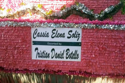One side of a parade float decorated with pink crepe paper and gold fringe. A sign posted on it reads: Cassia Elena Soliz Escorted by Trisian Daniel Botello.
