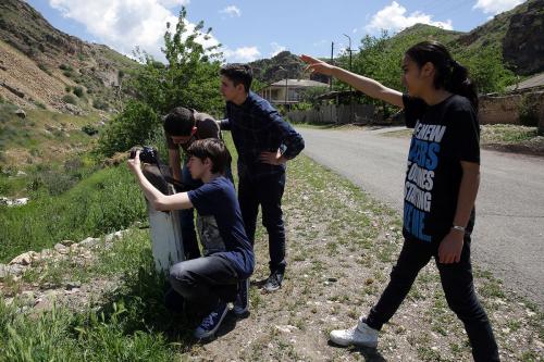 Four teens, three boys and a girl, look and point to something out of view. One boy is kneeling and aiming his camera in that direction.