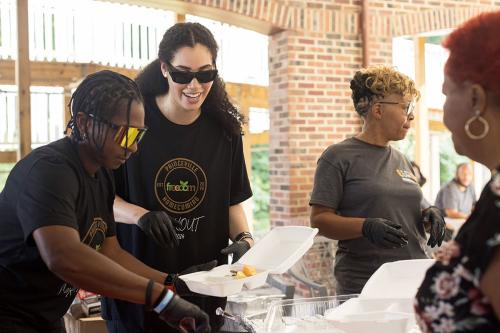 Three people wearing sunglasses and black safety gloves dish up food in to-go containers, while one person waits to be served.