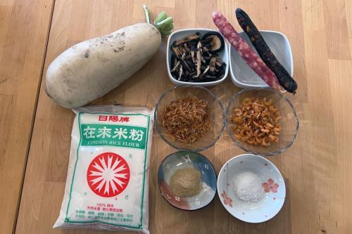 Ingredients arranged on a table: a plump white daikon radish, bag of rice flour, bowl of mushrooms, two skinny sausages, bowls of fried shallots and dried shrimp, and two bowls of powdery substance.