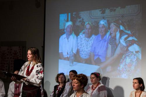 Holding a lyrics sheet, a woman with chunky red jewelry and white embroidered blouse sings. Behind her are other performers, seated, and a screen with a projection of elder woman around a table, all wearing head scarves.