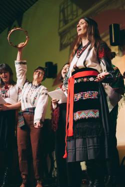 Five singers on stage in traditional embroidered outfits. One raises a tambourine high.