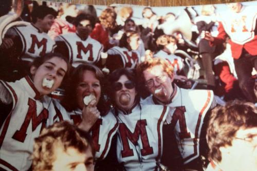 Older photo of band members in uniform. Four lean together in front, each holding a half-eaten apple in their mouth.