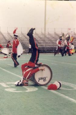 Older photo of a band member in full uniform, standing on their head on the field, playing the bass drum strapped to their chest which rests on the ground.