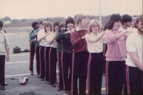 Older photo of band members in rehearsal dress, lined up and each massaging the shoulders of the person in front of them.