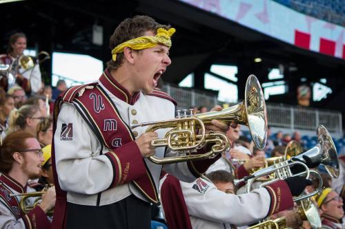 A young band member in uniform holds his mellophone (shaped like a tuba but much smaller) like a weapon, shouting from the stands in a stadium.