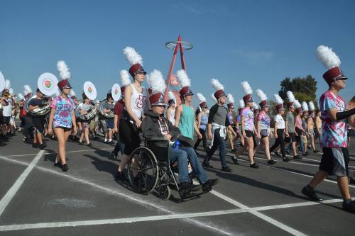 Band marching in formation in plain cloths except for hats with large white feathers sticking up. One member in the front is using a wheelchair, pushed by another member.
