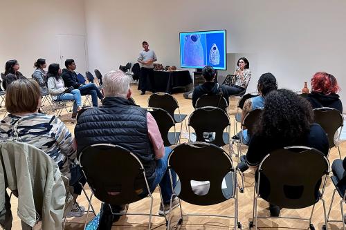 A crowd of people seated in a semicircle watch a man giving a presentation.