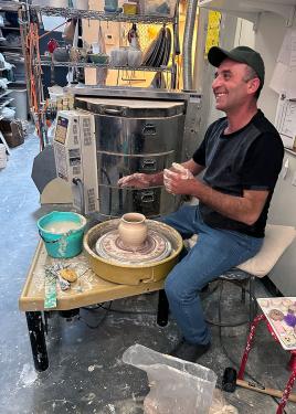 A man works at a pottery wheel, smiling.