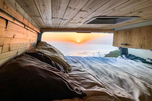 From inside a van, a bed and wood panelling in the foreground, and a flowing sunset over the ocean through the open back door.
