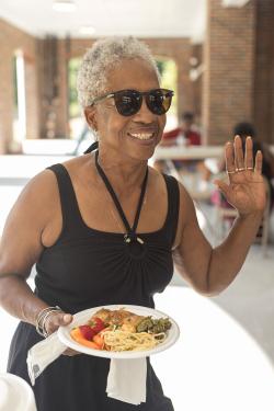An elder woman with short gray hair and a black tank tops waves with one hand and holds a plate of food in another.