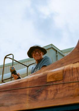 A man wearing glasses and bucket hat works with a tool on a wooden boat.