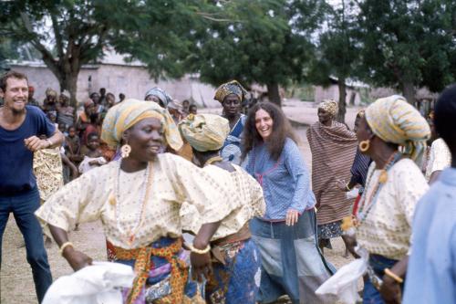 A group of mostly African women, and a white man and woman, dance together outdoors.