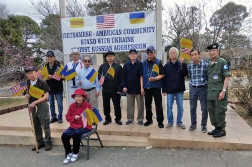 Several elder people pose in front of the stone monument, now holding a banner that reads Vietnamese-American Community stand with Ukraine against Russian Aggression. Most people hold three small flags: United States, South Vietnam, and blue and yellow Uk