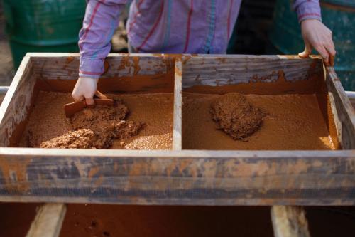 Close-up of a person scraping wet clay through a rectangular wooden sieve.