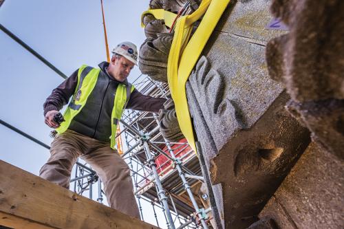Dressed in a white hard hat and bright green safety vest, Joe Alonso examines a piece of carved stone, held in place by a yellow strap. Behind him, towers of scaffolding and a clear blue sky.
