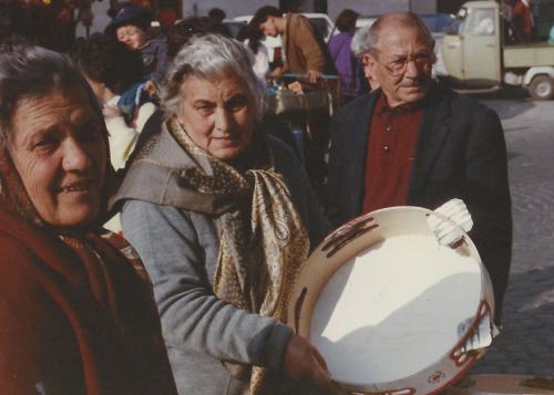An older woman holds a frame drum, flanked by two peole at her sides.