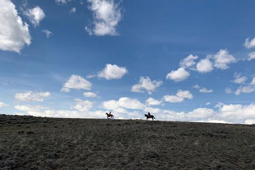 From afar, two people riding horseback along a grassy ridge, silhouetted by the blue sky and white clouds behind them.