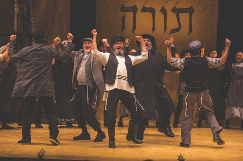 Theater production, men dancing on stage with Hebrew characters on the backdrop.