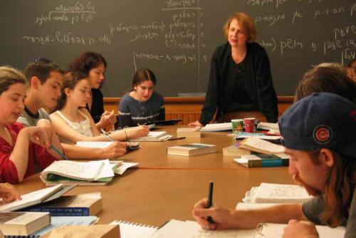 In a classroom, a teacher stands at the front of a table, surrounded by students with notebooks and books. Yiddish text is written on the blackboard behind her.