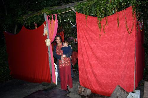A woman holds a baby, standing at the entrance of a temporary structure with walls of hung red fabric at night.