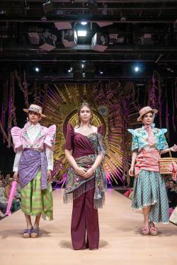 Three people pose on a runway stage. The two on the sides wear multicolored ensembles of button-up shirts, blouses with high pointy sleeves, wrap skirts, and straw hats. The woman in the center wears a maroon pant suit with high pointy sleeves and a flora
