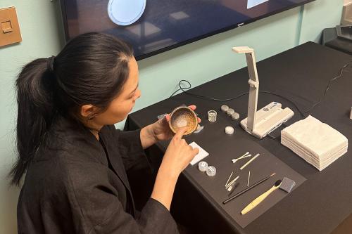A woman with long black hair in a ponytail, wearing all  black, sits at a black table, pointing to the inside of a small ceramic cup. On the table in front of her are small containers and tools including Q-tips, toothpicks, a toothbrush, and a paintbrush,