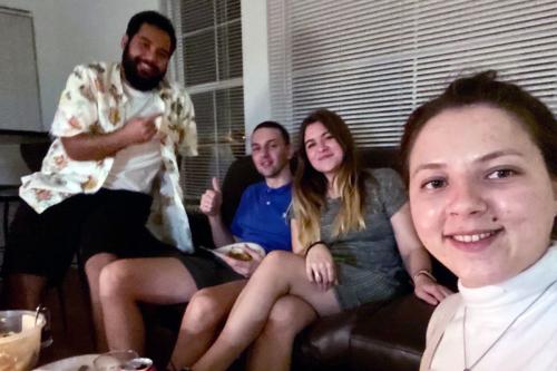 A selfie by a young woman, with three other people behind her sitting on a couch.