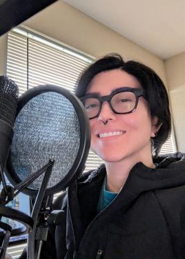 A person with short dark hair, black glasses, and black jacket smiles at the camera, standing in front of a studio microphone with a circular pop screen.