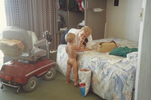 Old color photo of a naked toddler, facing away, feeding a potato chip to an older woman who is leaning over a bed. A mobility scooter is parked to the side.
