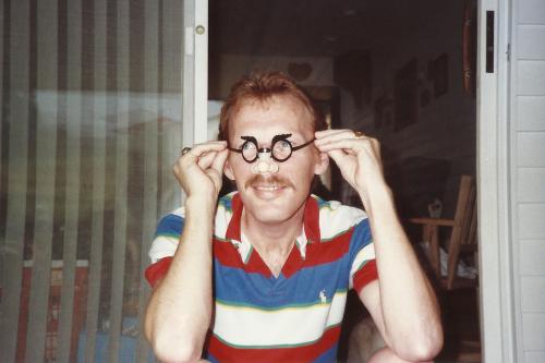 Old color photo of a man holding up a pair of novelty glasses to his face.