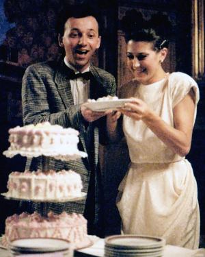A newlywed couple, still in suit and gown, holds a slice of wedding cake on a plate. The three-tiered cake is on the table in front of them.