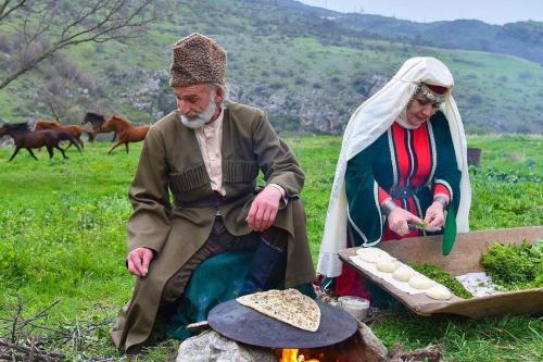 Seated outdoors among green grass, a man tends an open fire cooking a piece of flatbread. The woman next to him cuts green herbs. A herd of horses run by in the background.