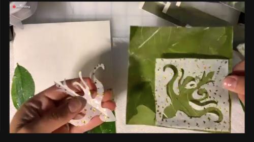 Zoom screenshot showing a closeup of hands showing pieces of cut paper, with the same bird design as the previous image.