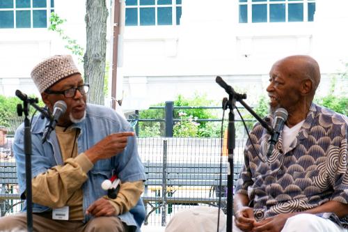 Two men sit on an outdoor stage in front of microphones. One of them speaks while pointing to the other, who is smiling back at him.