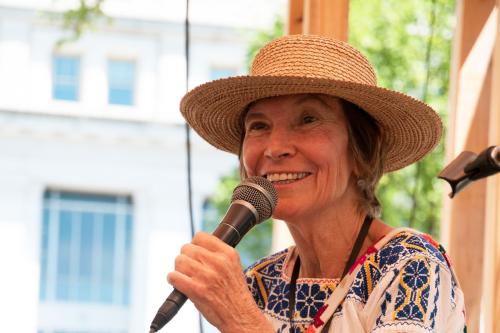 A woman with woven straw hat and embroidered blouse smiles, presenting on an outdoor stage with microphone in hand.
