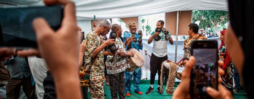 Four men stand together on a tented stage playing percussive instruments. They are dressed in brightly patterned clothes and are filmed by people holding cellphones.