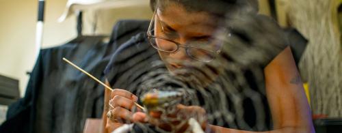 A young woman wearing glasses paints a doll she holds in her hands. In the foreground appears a spiderweb.