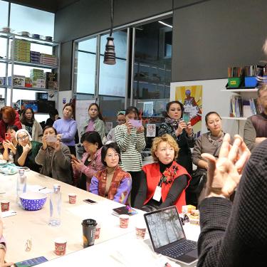 Twenty women sit attentively around a conference table facing a presenter with a laptop.