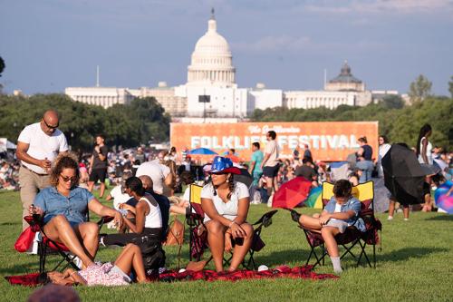 Crowds of people sit on the grass of the National Mall, with an orange Folklife Festival mural and the U.S. Capitol in the background.
