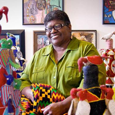 An African American woman poses and smiles among colorful artwork: paintings in the background, fabric sculptures in the foreground, and she is holding an African patterned textile.