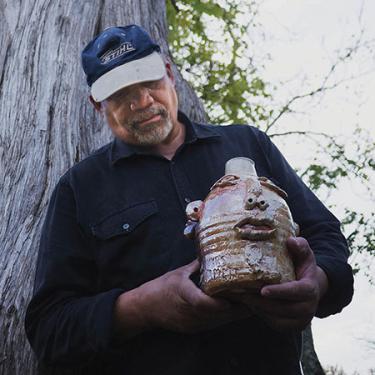 An older Black man holds up a ceramic jug shaped like a human face.