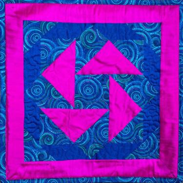 Quilt square containing four triangles and a square border in hot pink on a background cloth of blue-green swirls.