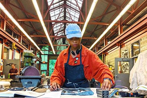 A woman reaches across a table while standing in a studio wearing a blue hat, an orange collared shirt, and a blue smock. Behind the woman are studio materials.