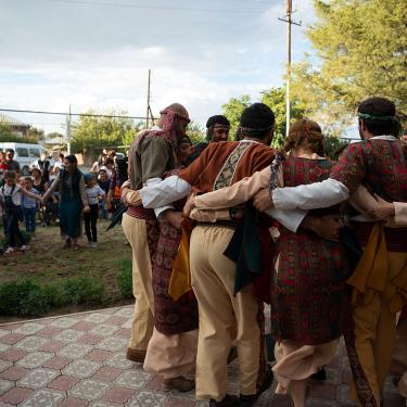 To the right, a group of adult men and women dance in a circle, hands clasped around each other's backs. To the left, young children also dance hand in hand.